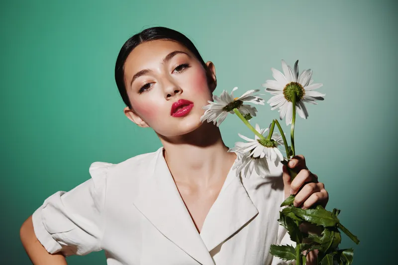 Young woman with red lips poses elegantly against teal background holding wilting white flowers. Fashion and beauty creative concept emphasizing drama and subtle melancholy mood.