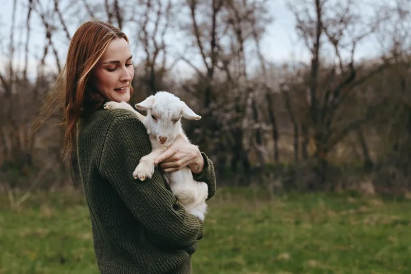 A woman in a cozy sweater holds a gentle goat kid outdoors in a sunlit field, sharing a warm moment. This scene conveys friendship, tenderness, and rural life with animals and nature.