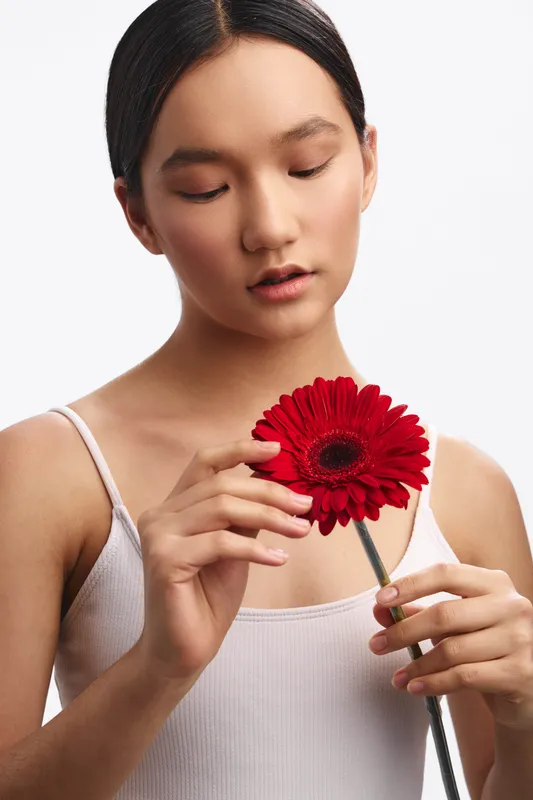 An asian woman in a light tank top gently holds a red flower, her gaze soft and thoughtful as she admires the bloom, conveying calm beauty and quiet celebration of nature.
