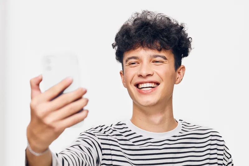 smiling man taking selfie with smartphone wearing striped shirt on white background showing happiness and casual style