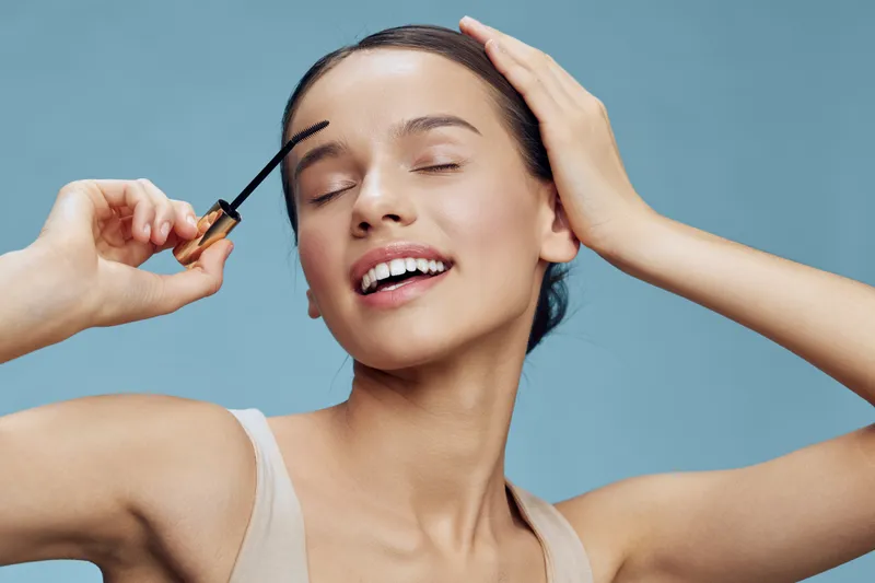 Young woman applying mascara with confidence, showcasing beauty and joy against a soft blue background, embodying self care and makeup techniques