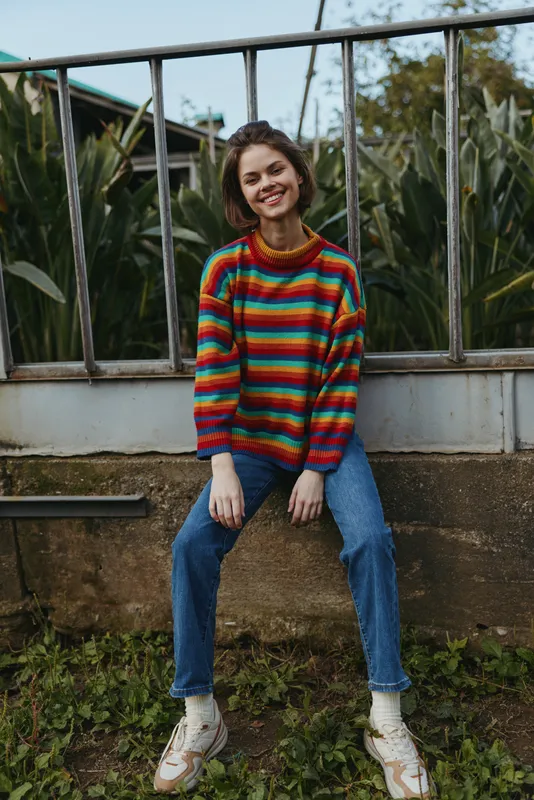 Woman smiling in a colorful striped sweater sitting on a garden wall, young adult early 20s in casual outfit with jeans and sneakers, relaxed pose and warm natural light for lifestyle fashion.