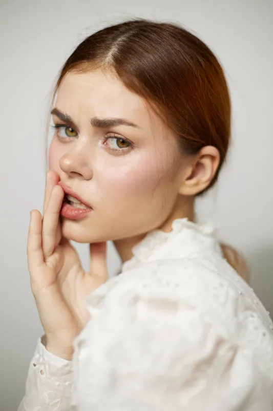 Young woman with expressive eyes in a white blouse, showing concern, with a soft background in neutral tones, reflecting a moment of vulnerability and emotion