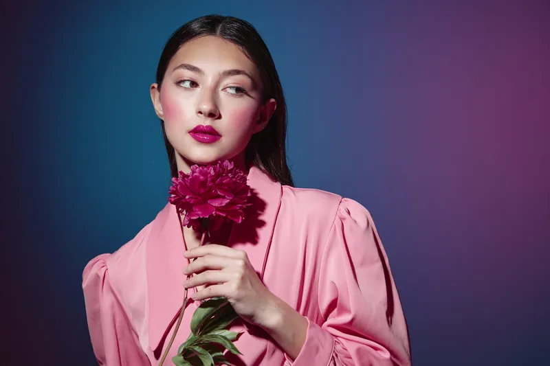 Fashionable woman in a pink outfit holds a burgundy flower while looking sideways. Studio portrait with contrast lighting in vibrant pink and dark blue colors. Art beauty concept.