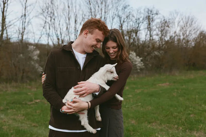 Young couple embraces outdoors while holding a white baby goat in a grassy field during early spring with bare trees in the background, sharing a tender moment together.