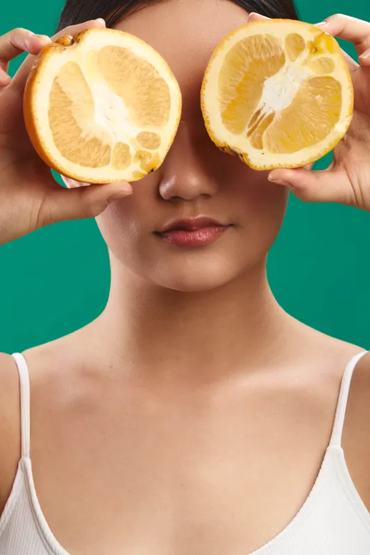 Asian woman playfully covers her eyes with lemon halves, presenting a fresh, vibrant portrait that blends wellness and citrus imagery in a bold studio setting.