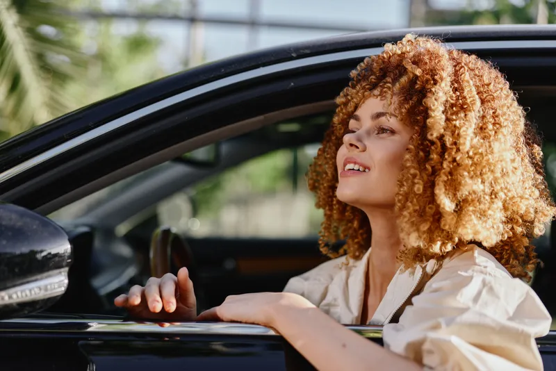 woman with curly hair sitting in car looking outside happily in sunny day, natural light, casual clothes, urban background
