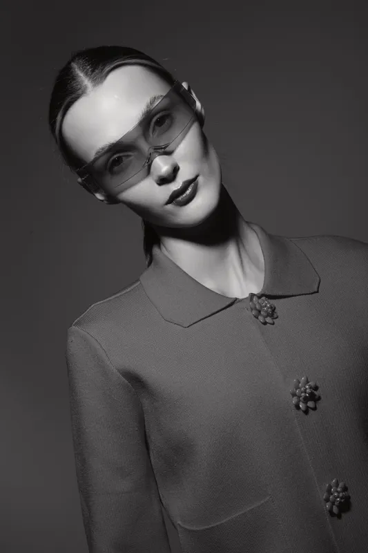 Young caucasian woman with pale skin wearing futuristic sunglasses and stylish jacket poses in studio with monochrome lighting, high fashion concept.