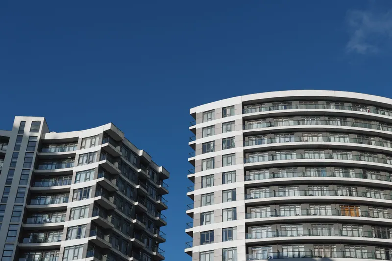 Sunny urban highrise with glass facade and balconies against a deep blue sky, showcasing modern architecture, vertical lines, and a clean cityscape with warm sunlight and symmetry.