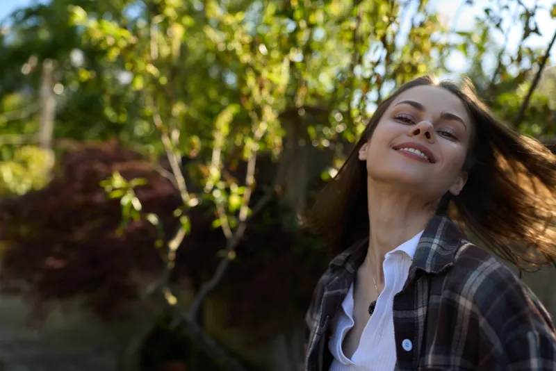Young woman in a plaid shirt smiles softly outdoors in a sunny garden, capturing a relaxed lifestyle moment with natural light and greenery nearby