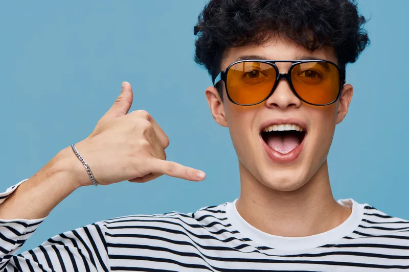 young man wearing orange sunglasses and striped shirt making call gesture with hand, expressing excitement against blue background
