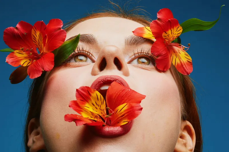 A vibrant portrait of a young woman expressing joy with colorful flowers in her mouth, showcasing beauty, nature, and creativity against a blue backdrop