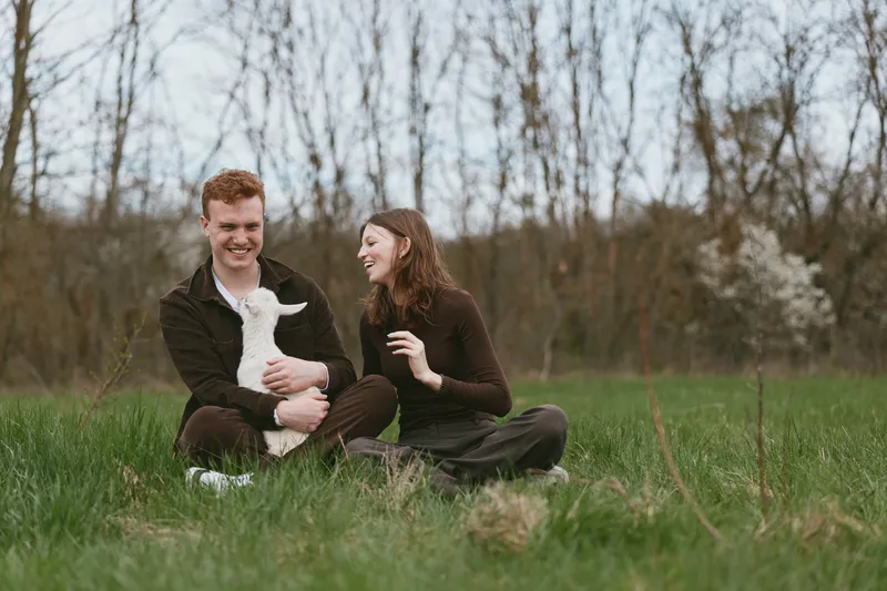 Cheerful couple outdoors with a playful goat kid, seated on grass, warm sunlight, relaxed mood, gentle breeze, natural tones, serene countryside backdrop, candid lifestyle moment