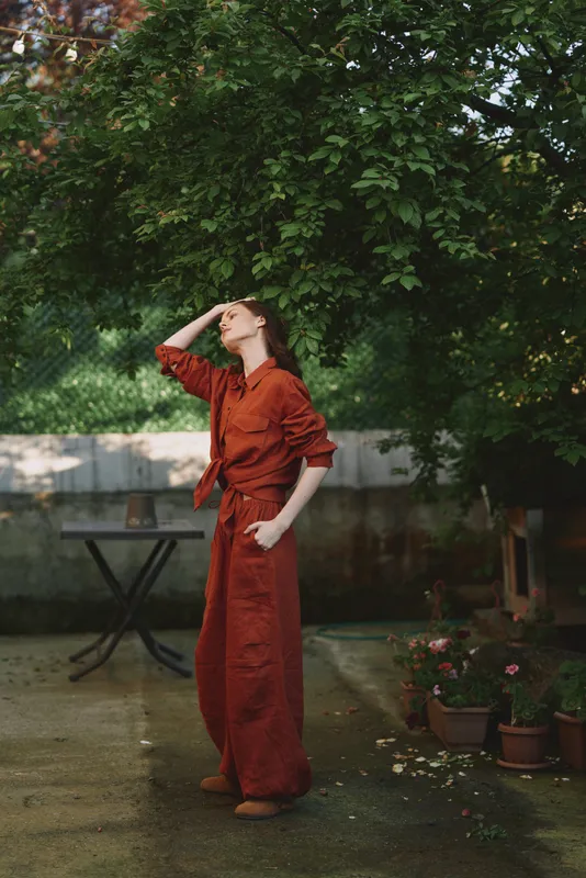 A stylish woman outdoors in a red dress pauses with a hand on her brow, sunlight filtering through trees. A candid lifestyle moment highlighting fashion, confidence and nature