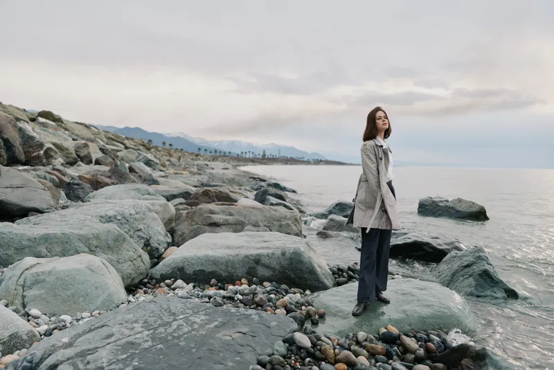 A woman in a trench coat stands on large rocks by the sea under a cloudy sky. The coastal landscape with pebble stones and distant hills creates a calm, contemplative mood.