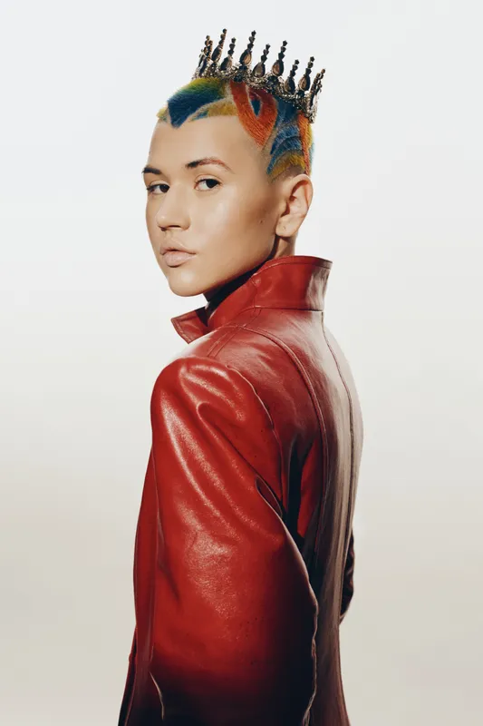 young woman with colorful short hair wearing stylish red leather jacket and unique crown accessory, posing confidently against plain white background