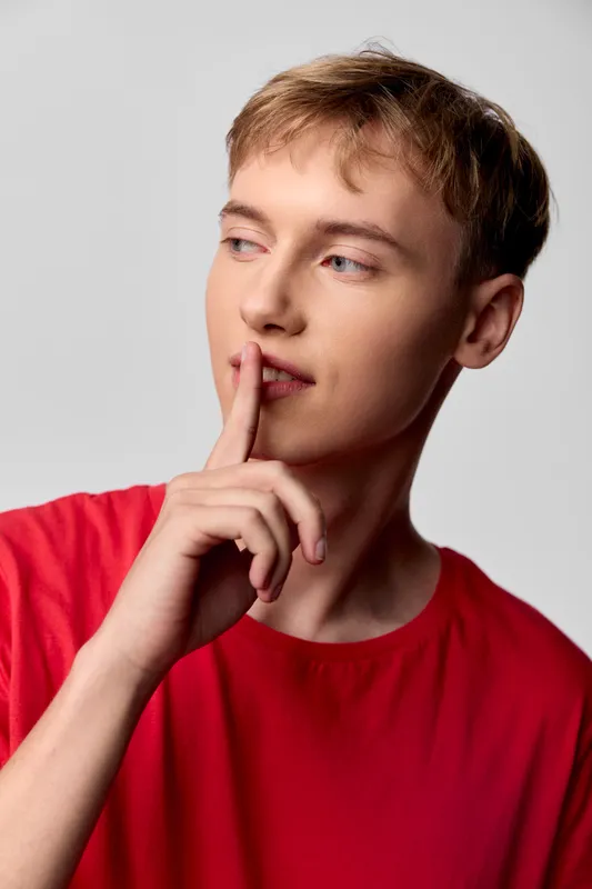 Thinking young man in a red shirt with finger to lips, pensive mood and calm gaze, studio setting conveys contemplation, focus, and a quiet moment of decision making