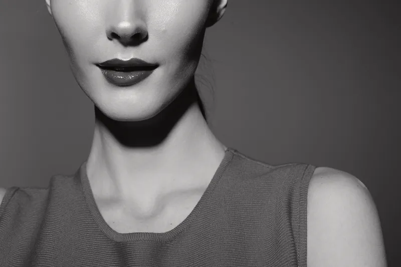 Close-up of a young white woman with pale skin and red lips wearing a sleeveless top. The monochrome portrait highlights elegance, beauty, and fashion concepts in studio lighting.