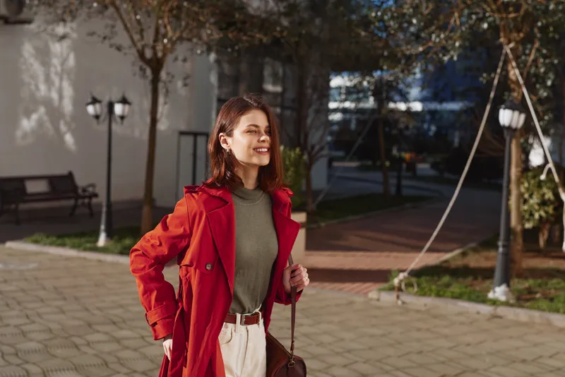 Young woman in a red coat smiles outdoors in a sunlit courtyard, green sweater, beige pants, carrying a bag, relaxed pose amid urban trees.