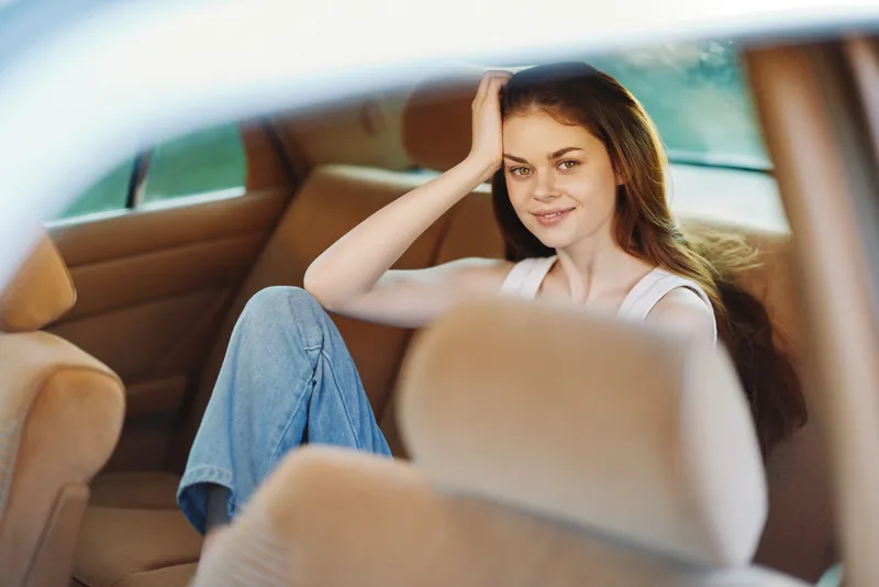 Smiling young woman with long hair sitting in a car, wearing casual clothes, enjoying a moment of tranquility in a bright environment.