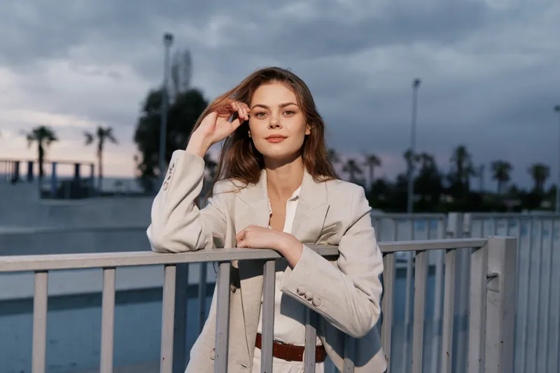 Businesswoman posing confidently in a stylish blazer against a dramatic sunset background, showcasing professionalism and grace in a modern setting.