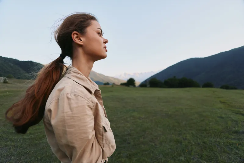 Young woman with long brown hair stands thoughtfully in an open green field surrounded by mountains under a clear sky.