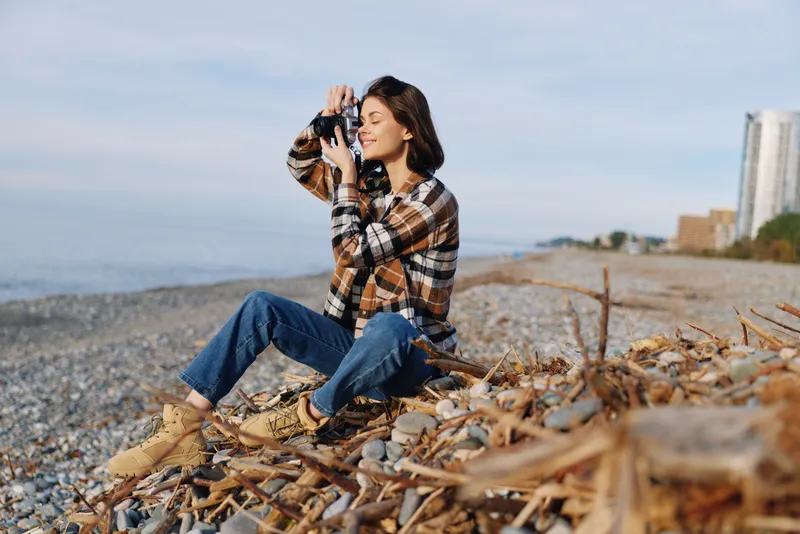 Photography woman on a beach with a camera in hand exploring the coast and sea breeze in casual outfit capturing candid scenery and warm light