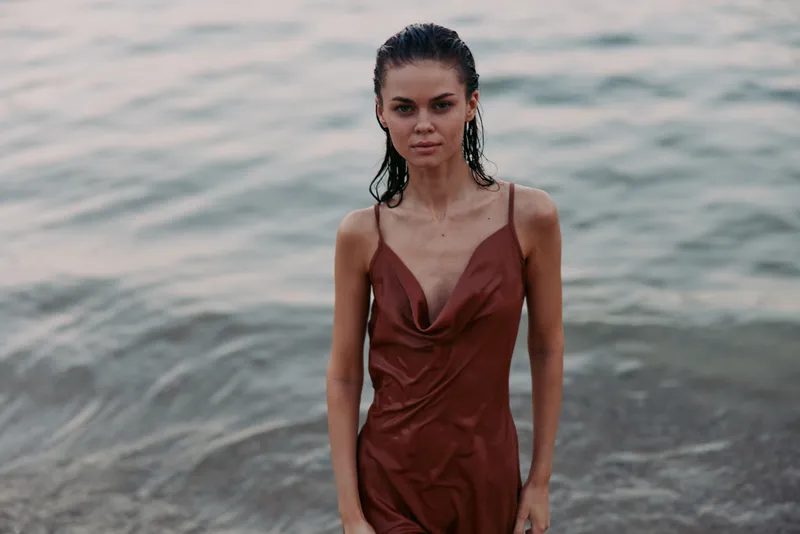 Portrait of a young woman standing near a calm shoreline in a sleeveless brown dress, soft light, poised expression, and graceful presence by the sea.