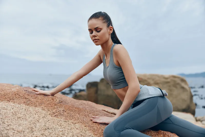 Fit young woman in activewear relaxing on the rocky beach, showcasing a healthy lifestyle and mindfulness in a serene outdoor setting