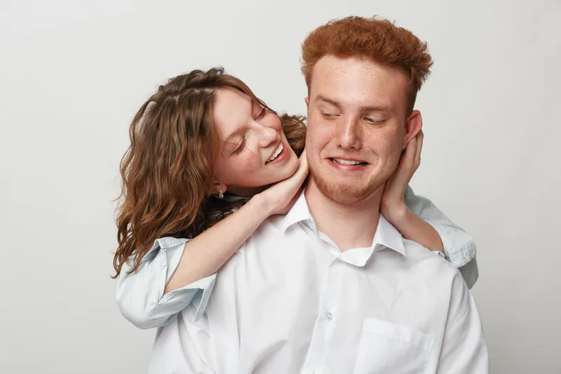 Happy young couple in casual white shirts share a playful moment. Woman embraces man from behind with smile, expressing affection and joy in a bright studio setting.