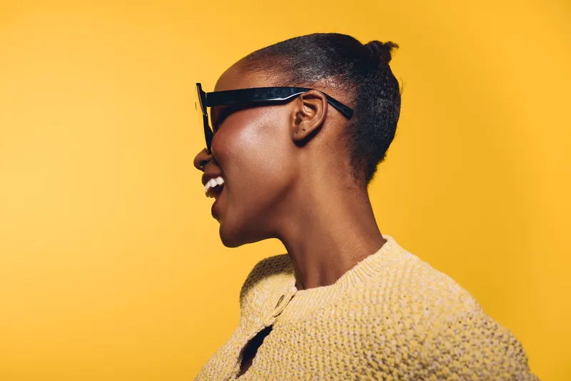 Profile of a smiling African woman with dark skin and sunglasses wearing a yellow knitted sweater against a bright yellow background. Concept of joy, fashion, and lifestyle.