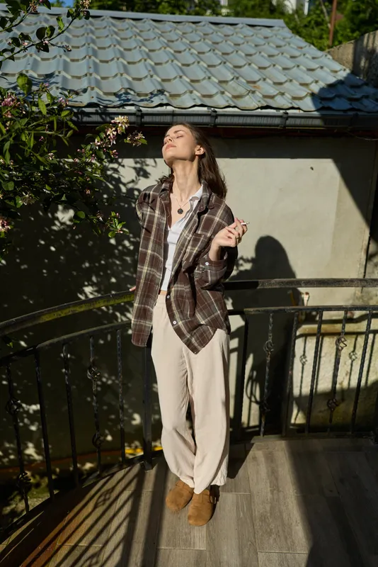 Sunlit woman on a balcony wears a plaid jacket and wide beige pants, standing barefoot, savoring warm sunlight and a relaxed, confident mood in a casual street style moment
