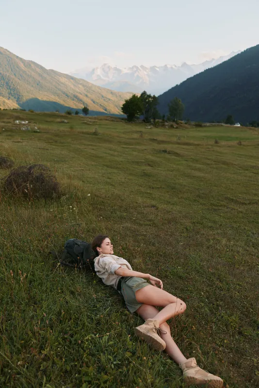 Young woman relaxing on grassy hill outdoors, enjoying peaceful nature, casual outfit, summer day, mountains in background, serene lifestyle moment