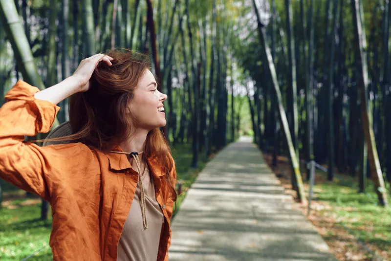 Woman smile redhead in orange jacket walking on a bamboo path through a sunlit forest, looking sideways. Outdoor portrait, natural light, peaceful travel and leisure scene.