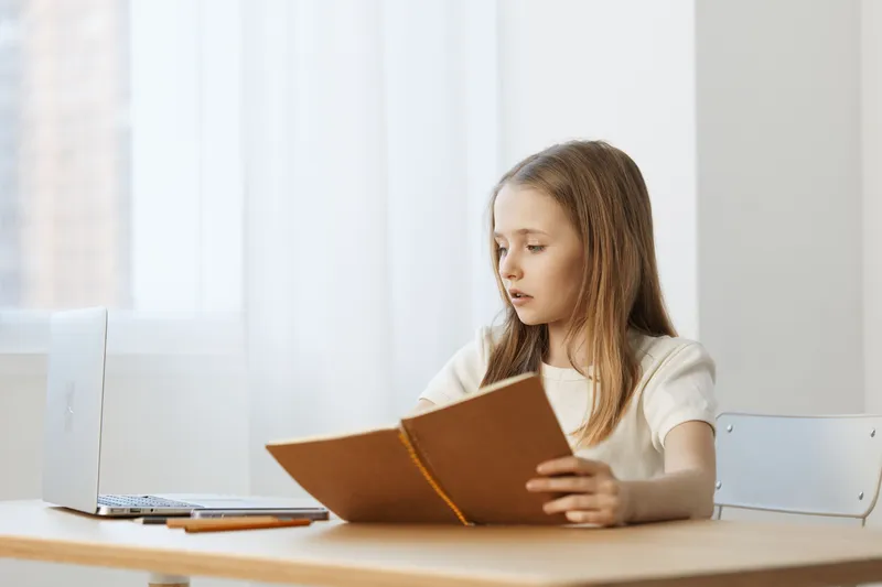 Young girl studying at a desk with a laptop, focused and thoughtful, surrounded by bright natural light and a minimalistic white background