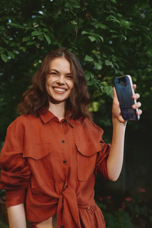 Smiling brunette woman with wavy hair holds up a smartphone outdoors, captured in warm natural light, conveying a casual selfie moment