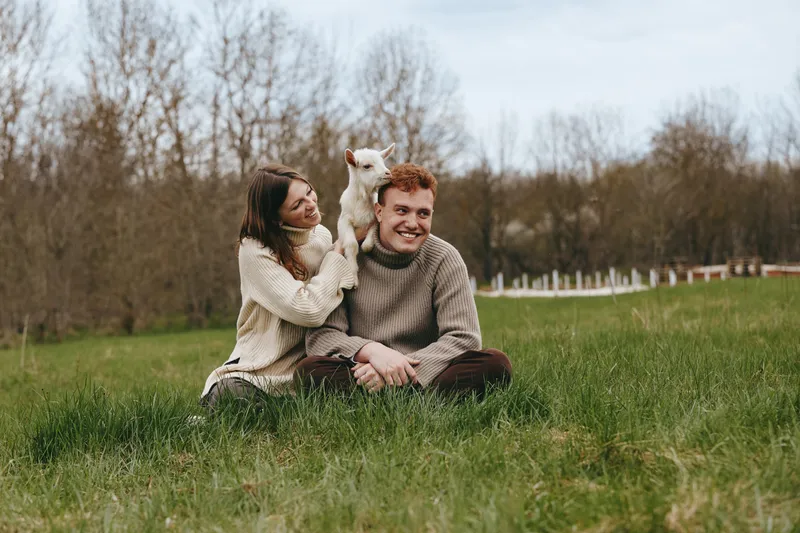 Smiling couple outdoors on a grassy field with a playful goat kid, sharing a tender moment. Natural light, candid lifestyle scene, casual attire, peaceful rural setting, warm atmosphere, connection