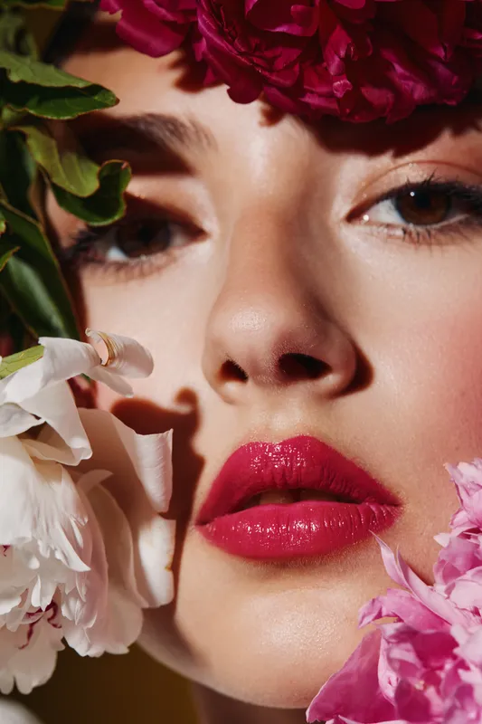 Close-up of a young woman with vibrant pink lips surrounded by colorful flowers. Crisp skin details and floral accents create a stylish beauty and fashion concept.