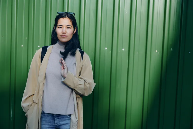 Woman in beige coat and gray sweater stands before a green metal wall, casual outdoor street fashion, confident pose, urban vibe, candid modern style, daytime scene