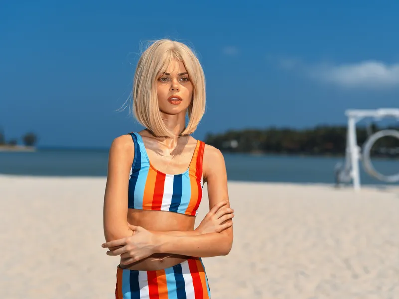 Blonde woman in colorful striped swimsuit standing on sunny beach with blue sky, summer vibe, confident pose, warm light, vacation mood.