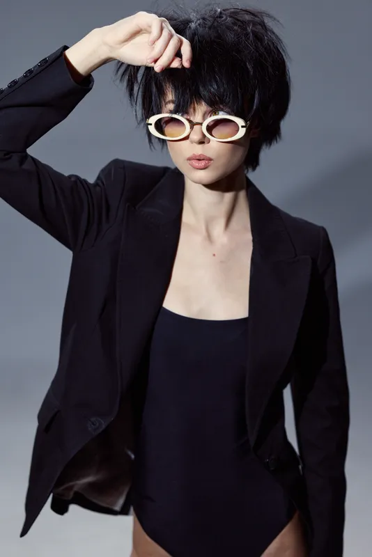 Stylish woman with short hair wearing white sunglasses and black blazer posing confidently in studio lighting