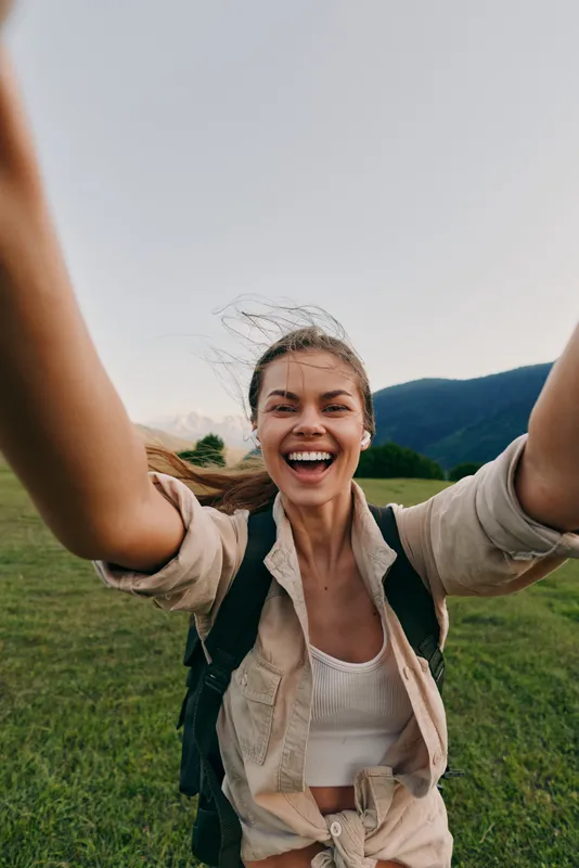 Young woman with long hair takes a joyful selfie outdoors in a green field with mountains in the background, wearing a beige jacket and white top on a clear day.