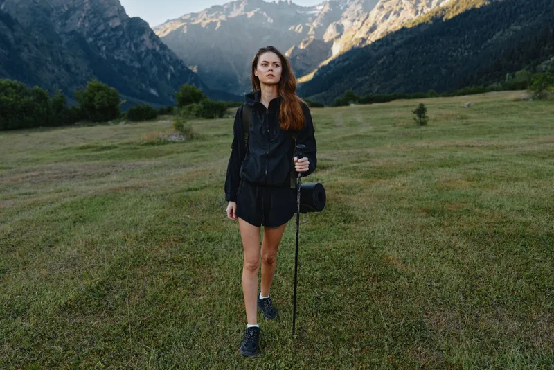 A solo hiker stands in a grassy meadow with distant mountains behind her. She wears a dark jacket and shorts, holding a trekking pole, ready for an outdoor adventure and scenic excursion.