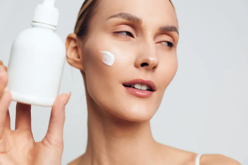 Woman applying white face cream from a bottle, closeup on smooth skin, clean white studio background, soft light, calm expression, concept of skincare, beauty, wellness, hydration, self care