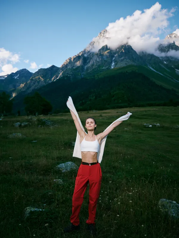 Athlete in red pants and white top stands in a wide alpine meadow with raised arms, mountains and blue sky behind, conveying freedom, energy and outdoor wellness