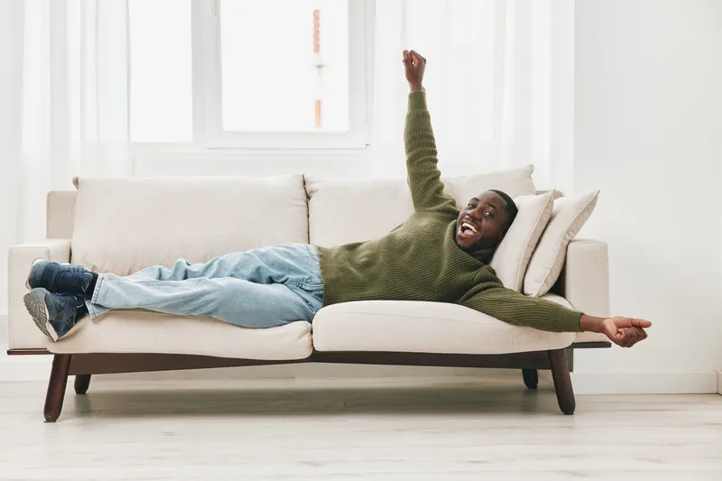 A joyful young Black male lounging on a modern couch, expressing happiness in a cozy, bright living room with light colors and natural light