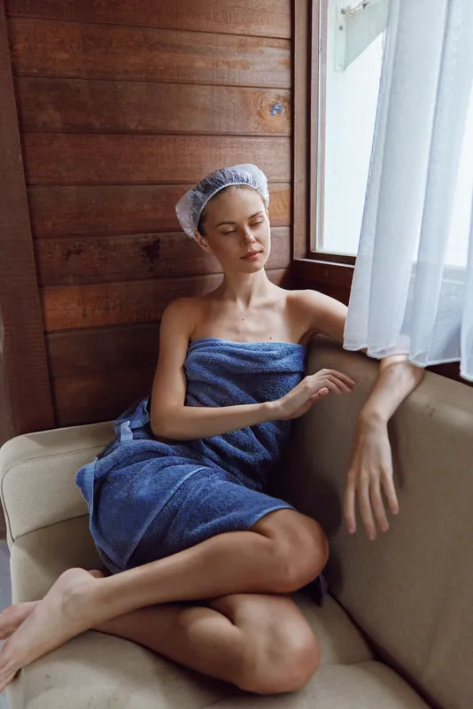 Relaxed woman wrapped in blue towel with shower cap sitting on beige sofa by wooden wall and white curtain, enjoying peaceful moment in natural light interior.