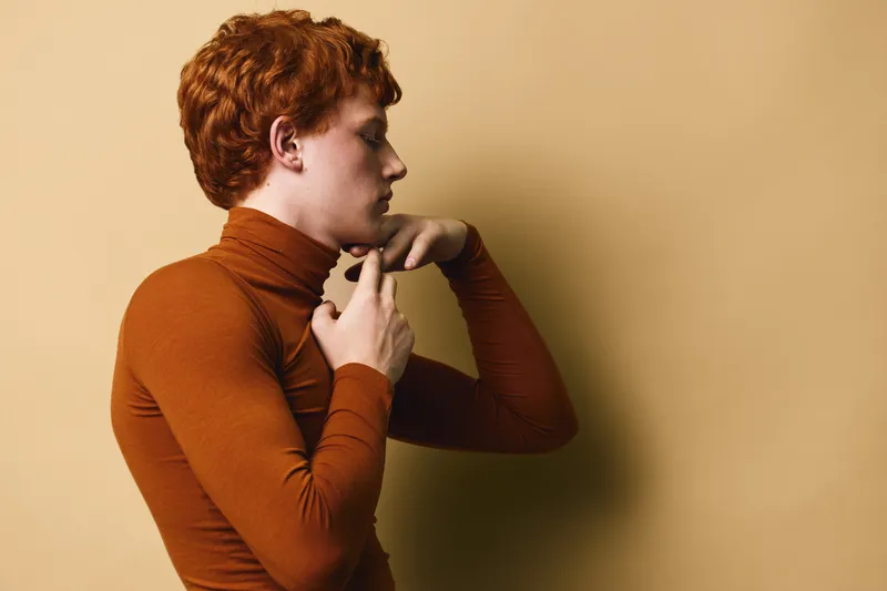 Young redhead person adjusting turtleneck sweater against beige background, side profile portrait with casual style and minimalist warm color tones.