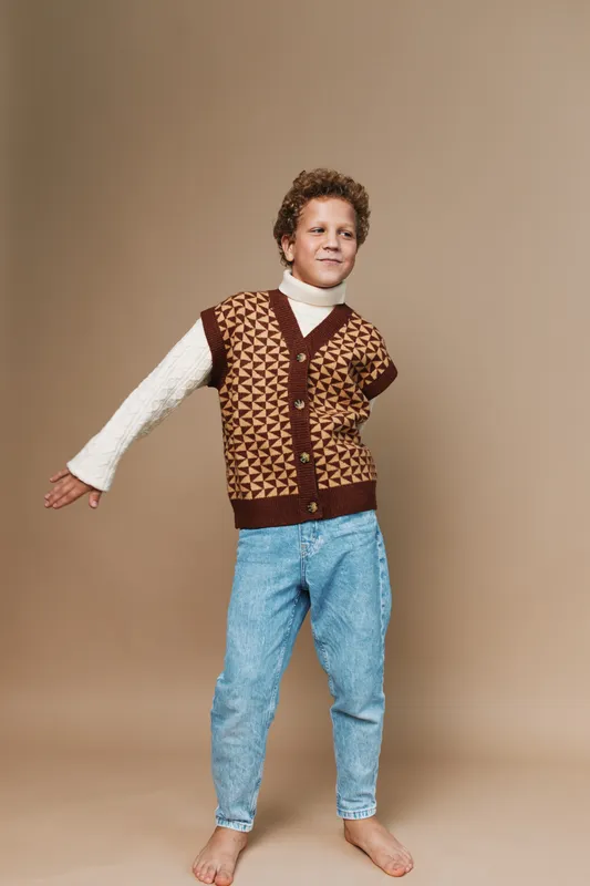 A playful young boy stands barefoot in a vintage geometric patterned cardigan over a white turtleneck, posing with one arm extended against a warm neutral backdrop.