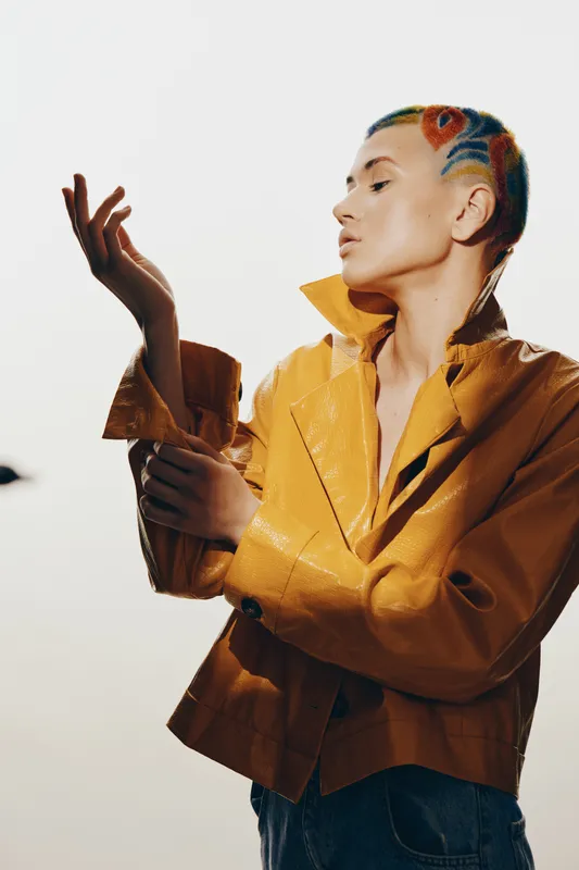 Artistic woman with colorful braided hair wearing a mustard yellow jacket poses thoughtfully against a bright minimalistic background, showcasing unique fashion and style.